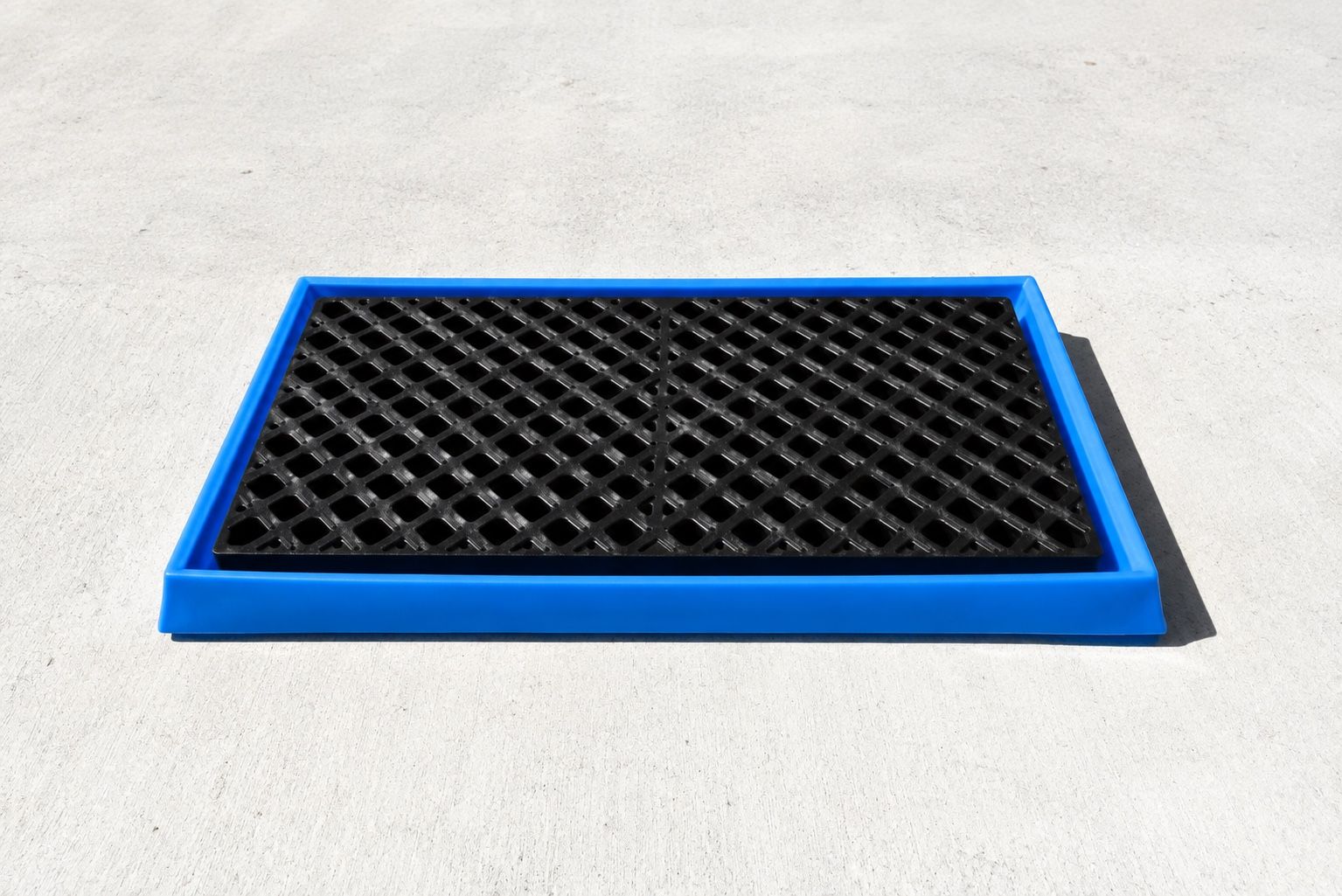 Blue rigid containment tray on a concrete floor with a black HDPE diamond-pattern grating panel lying inside it, the tray wall standing clear around the edge of the grating