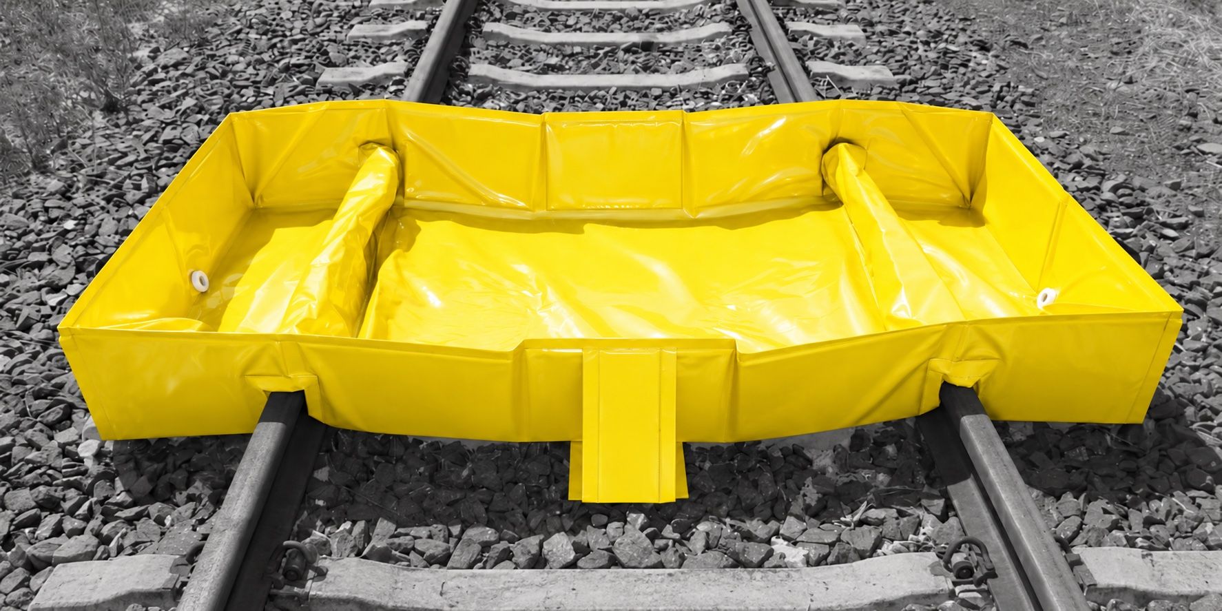 Yellow PVC rail spill bund sitting between two rails, its front and back walls notched down over each rail, with diagonal side supports inside the corners and drain fittings in the side walls